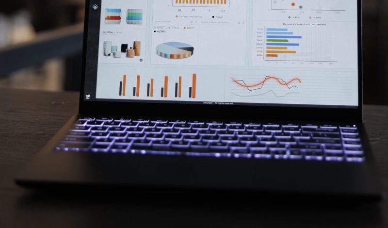 Laptop computer with marketing statistics graphs on screen, standing on table in startup office, coworkers discussing company strategy in background. Employees working overtime in startup office